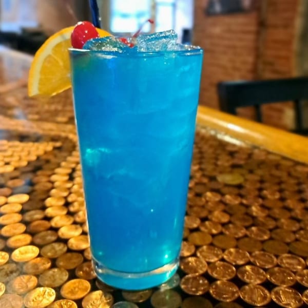 A vibrant blue cocktail with ice, an orange slice, and cherries sits on a penny-covered bar.