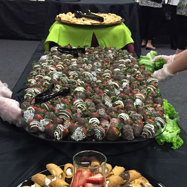 A dessert buffet with chocolate-covered strawberries, cannolis, and a pie, with hands in gloves.