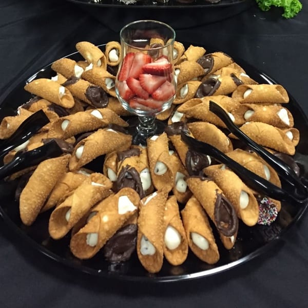 A black platter filled with cannolis, some dipped in chocolate, surrounding a glass of sliced strawberries.