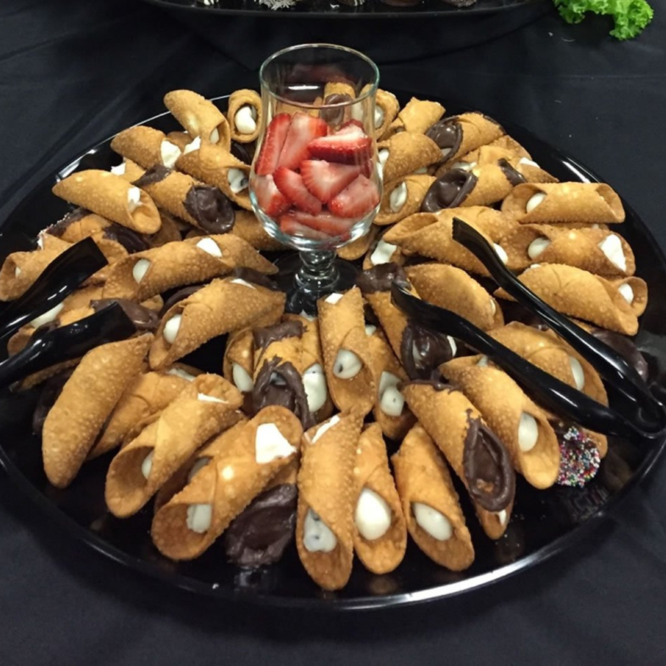 A black platter filled with cannolis, some dipped in chocolate, surrounding a glass of sliced strawberries.