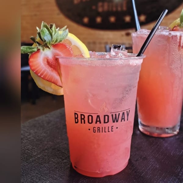 Two pink cocktails with ice, garnished with strawberries and lemon slices, on a dark table.