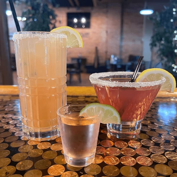 Three cocktails, including a tall drink, a martini, and a shot, on a penny-covered bar.