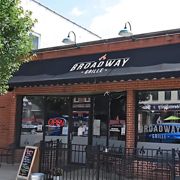 Exterior of Broadway Grille, a brick building with a black awning, outdoor seating, and an "OPEN" sign.