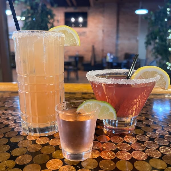 Two cocktails and a shot glass with citrus garnishes on a penny-covered bar top.