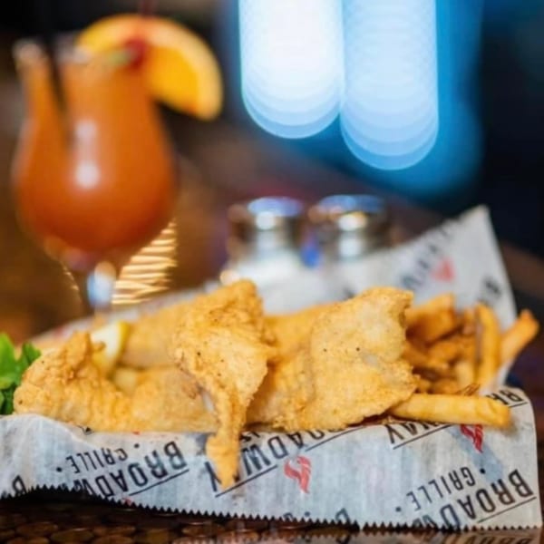 Fried fish and french fries in a basket with a cocktail and salt and pepper shakers.