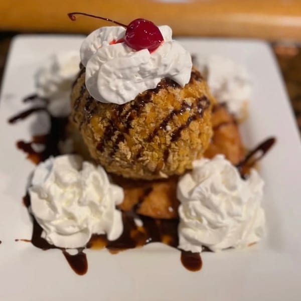 Fried ice cream dessert with whipped cream, chocolate syrup, and a cherry on a white plate.